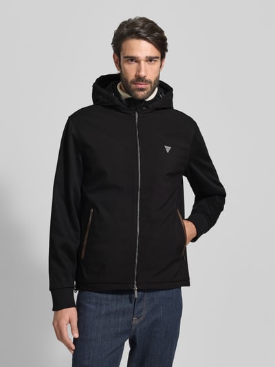 Männermodel trägt Windbreaker mit Zweiwege-Reißverschluss von Guess in Schwarz, Regular Fit, Kapuze, Reißverschlusstaschen, Label-Detail, Ganzkörperansicht von vorne.