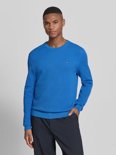 Mannenmodel draagt Regular Fit Strickpullover aus reiner Baumwolle van Tommy Hilfiger in effen blauw met gebreide look, long sleeve, logodetail, ronde hals, geribde mouwboord en rompzoom, puur katoen, voorkant halve lichaamsopname.