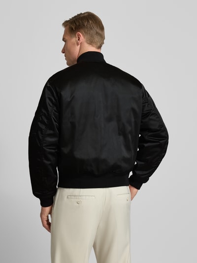 Männermodel trägt Regular Fit Bomberjacke aus Satin von Lacoste in Schwarz mit geripptem Abschluss, wasserabweisend, Ganzkörperansicht Rückansicht.