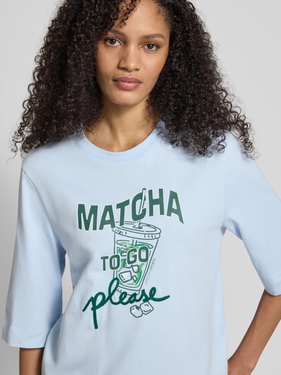 Kobiece modelka nosi Slim Fit T-Shirt mit Motiv-Print von Marc Cain z bawełny, z nadrukiem logo, półrękaw, dekolt okrągły, długość pleców 58 cm, jasnoniebieski, widok na górną część ciała z przodu.