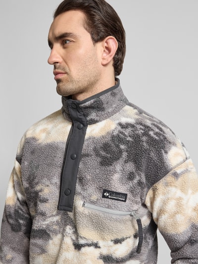 Męski model nosi Regular Fit Pullover mit Logo-Patch od Columbia, fließ-Optik, stójka z listwą z zatrzaskami, naszywka z logo, kieszeń na piersi zapinana na zamek błyskawiczny, elastyczne wykończenia, długość tyłu w rozmiarze M: 72 cm, długość rękawów w rozmiarze M: 49 cm, jasnoszaro-beżowe barwy, miękka tekstura, efekt melanżu. Nahaufnahme Oberkörper.
