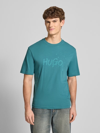 Männermodel trägt Regular Fit T-Shirt aus reiner Baumwolle Modell 'NIEROFIX_U261' von HUGO in Türkis mit Motiv-Print und Label Stitching, Rundhals, kurzarm, unifarben, Ganzkörperansicht von vorne.