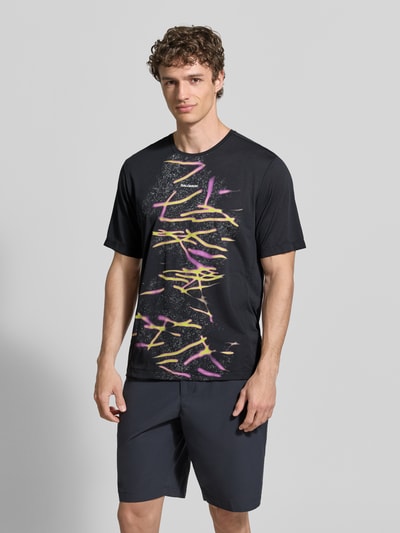 Männermodel trägt Herren T-Shirt von Salomon, gerade geschnitten, schwarzer Stoff, bunter Logo- und Motiv-Print, Modell 'Kout', Ganzkörperansicht von vorne.