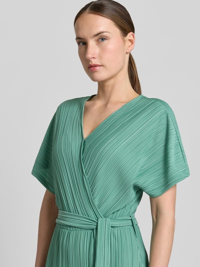 Vrouwenmodel draagt groene jumpsuit met plissévouwen, wikkellook, V-hals en strikceintuur, Pe44arl van ZABAIONE, detailopname bovenlichaam.