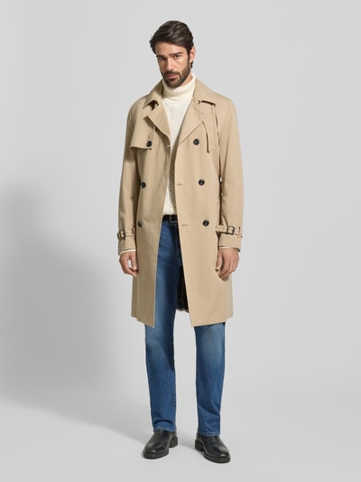 Männermodel draagt Regular Fit Trenchcoat met Gun Flap Model 'MALUKS2511' van HUGO, effen beige, tailleriem en loopsplit, lange mouw, steekzakken, binnenzak, waterafstotend, platte kraag, dubbele knoopsluiting, labelstitching, achterpas, geheel van voren te zien.