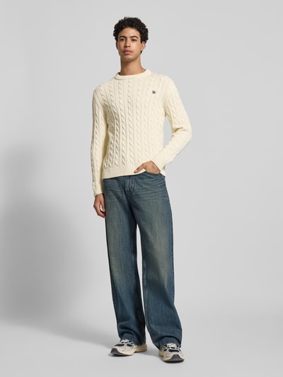 Männermodel trägt Slim Fit Pullover in Strick-Optik von Superdry aus reiner Baumwolle, Logo-Stitching, Rundhalsausschnitt, elastische Abschlüsse, helles Beige, grobe Textur, Ganzkörperansicht von vorne.
