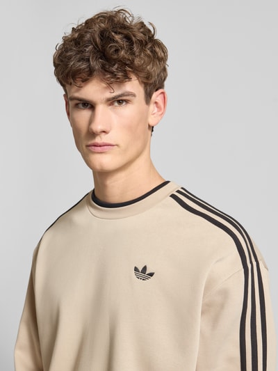 Męski model nosi Sweatshirt mit Label-Stitching od adidas Originals, mieszanka bawełny, krój regular fit, prążkowane wykończenia, okrągły dekolt, wyhaftowane logo, długość tyłu 69 cm, długość rękawów 60 cm, beżowy z czarnymi detalami, gładka tekstura, Nahaufnahme górnej części ciała.
