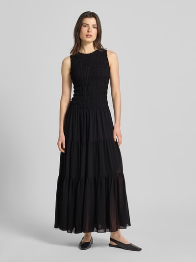 Frauenmodel trägt Maxikleid mit Smok-Details und Volantsaum von MICHAEL Michael Kors in Schwarz, Taillierter Schnitt, Rundhalsausschnitt, kurzer Reißverschluss am Träger, Rückenlänge 141 cm, Brustweite 31 cm, Ganzkörperansicht von vorne.