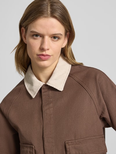 Kobiece model prezentuje kurtkę jeansową Regular Fit Jeansjacke aus reiner Baumwolle Modell 'BETT' von Pieces, jednokolorowy brąz, obszycie ze sztruksu na wykładanym kołnierzu, kieszenie z patką, denimowe wykończenie z czystej bawełny, wyraźna struktura materiału, zapięcie na zakrytą listwę z zatrzaskami, raglanowe rękawy, detaliczna faktura tkaniny, zbliżenie na górną część ciała.