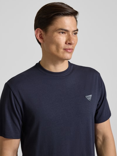Männermodel trägt T-Shirt mit Logo und Rundhalsausschnitt von Guess in Dunkelblau, Baumwoll-Mix mit Modal-Anteil, Relaxed-Fit, Nahaufnahme Oberkörper.