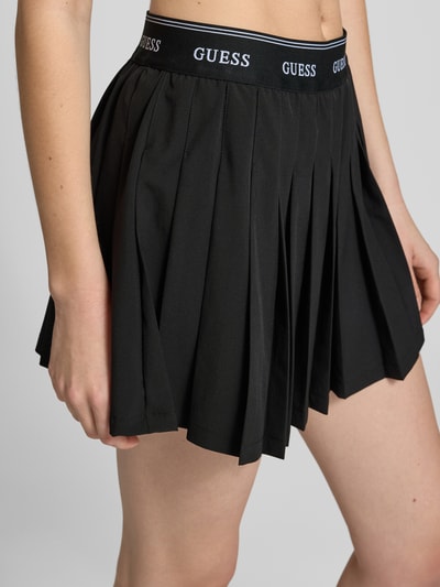 Frauenmodel trägt Faltenrock mit elastischem Bund Modell 'SELENE' von Guess Activewear, Minirock in Schwarz, Material-Mix mit Stretch-Anteil, A-Linie, Nahaufnahme Oberkörper. Bundweite 35 cm, Gesamtlänge 36 cm bei Größe S.