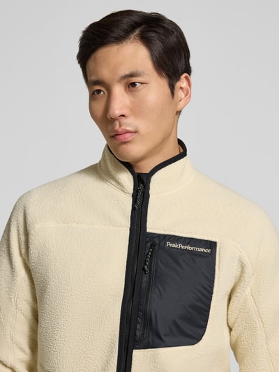 Mężczyzna nosi Jacke mit Teddyfell von PEAK PERFORMANCE, beżowa z czarną kieszenią, prosty krój, logo, zbliżenie na górną część ciała.