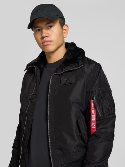 Männermodel trägt Bomberjacke mit Label-Patch und Kentkragen von Alpha Industries in Schwarz, Reißverschluss, Teddyfellkragen, Nahaufnahme Oberkörper.