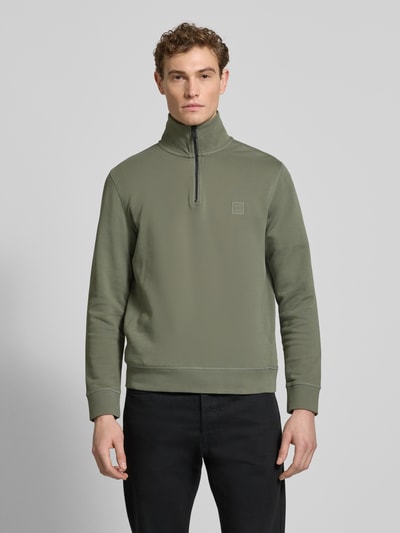 Męski model nosi Regular Fit Troyer aus reiner Baumwolle Modell 'TERRY' od BOSS Orange, gładki, krój regular fit, patch z logo, długi rękaw, elastyczne wykończenia, materiał dresowy z czystej bawełny, kolor oliwkowy; widok na całe ciało z przodu.