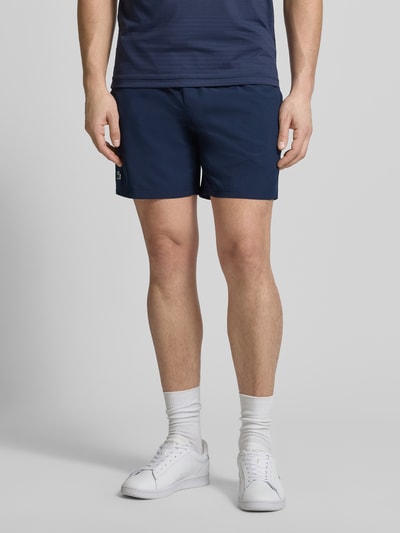 Männermodel trägt Loose Fit Sport-Shorts mit Mesh-Bund von Lacoste Sport, dunkelblau, unifarben, Logo-Stitching, Ganzkörperansicht von vorne.