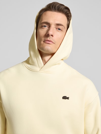 Männermodel trägt Regular Fit Hoodie aus reiner Baumwolle von Lacoste, efekt: jednolity kolor, naszywka z logo, dzianina dresowa, bawełna, bluza z kapturem, krój regular fit, długi rękaw, zakończenie rękawów: prążkowane, kieszeń kangurka, kolor jasny, nahaufnahme oberkörper.