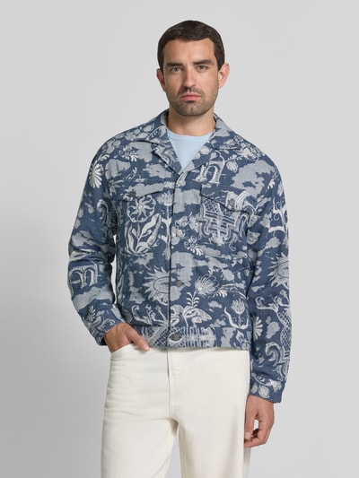 Męski model nosi Jacke im Allover-Look mit Reverskragen von Scotch & Soda, krój regular fit, wzór na całej powierzchni, listwa guzikowa, kieszenie na piersi, długość tyłu w rozmiarze M 61 cm, szerokość ramion w rozmiarze M 43 cm, szerokość klatki piersiowej w rozmiarze M 51 cm, długość rękawów w rozmiarze M 62 cm, odcienie niebieskiego i bieli, wyrazista tekstura, Ganzkörperansicht von vorne.