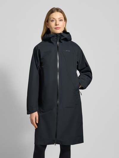 Frauenmodel trägt Mantel mit Zweiwege-Reißverschluss und Kapuze von PEAK PERFORMANCE in Schwarz, glatte Oberfläche, Ganzkörperansicht von vorne. Relaxed Fit, Kapuze, Logo vorn, Rückenlänge 101 cm, Schulterbreite 44 cm, Brustweite 53 cm, Ärmellänge 58 cm.