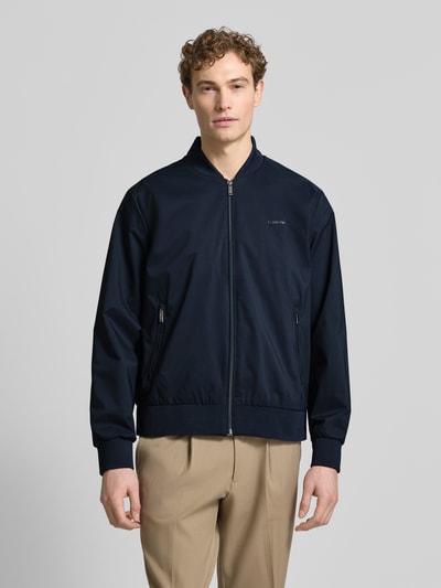 Männermodel trägt dunkelblaue Bomberjacke mit Reißverschlusstaschen Modell 'MATTE' von CK Calvin Klein, glatte Textur, Ganzkörperansicht von vorne.