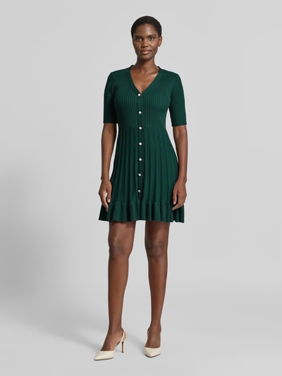 Frauenmodel trägt Strickkleid in Minilänge mit Viskose-Anteil von Christian Berg Woman in Dunkelgrün, Ripp-Optik, Rüschensaum, Knopfleiste, taillierte Passform, kurzärmlig, Ganzkörperansicht von vorne.