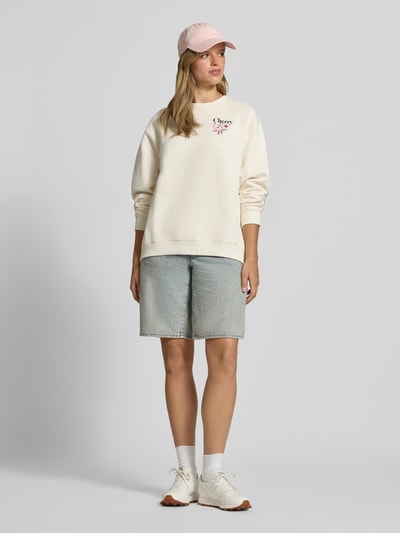 Modelka nosi bluzę Relaxed Fit Sweatshirt aus reiner Baumwolle Modell 'Classic Crew_B_4' od HUGO Blue w jasnym kremowym kolorze, luźny krój, gładka tekstura, z przodu widoczne różowe logo, do tego jasnoniebieskie szorty dżinsowe, białe sneakersy i jasnoróżowa czapka z daszkiem, Ganzkörperansicht von vorne.