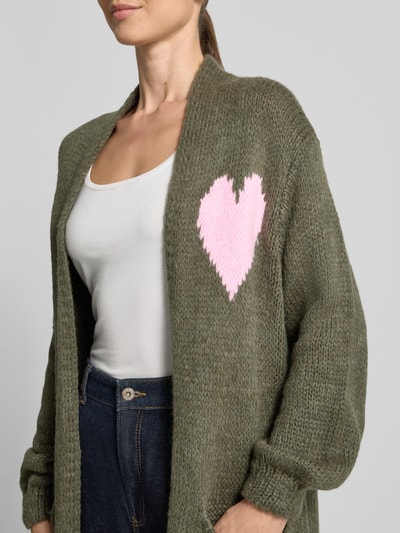 Frauenmodel trägt Strickjacke mit Woll-Anteil von miss goodlife, olivgrün mit rosa Herz-Motiv, lockere Passform, Nahaufnahme Oberkörper. Kontext: Material-Mix, offenes Design, aufgesetzte Eingrifftaschen, Rückenlänge 118 cm, Ärmellänge 55 cm.
