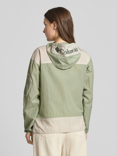 Frauenmodel trägt Regular Fit Windbreaker mit Logo-Print Modell 'Challenger' von Columbia in Hellgrün und Beige, Rückansicht, Kontext: Damenjacke mit Omni-Shield™ Technologie, Känguru-Tasche, Ärmeltasche, Kapuze mit Tunnelzug, elastische Abschlüsse, Maße Größe S.