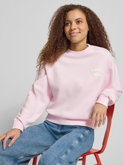 Frauenmodel trägt Boxy Fit Sweatshirt aus reiner Baumwolle von Tommy Jeans in Optik: Unifarben, Label-Print, Crew Neck, Langarm, gerippte Abschlüsse, Sweat-Material, zart rosa, glatte Struktur, Ganzkörperansicht von vorne.