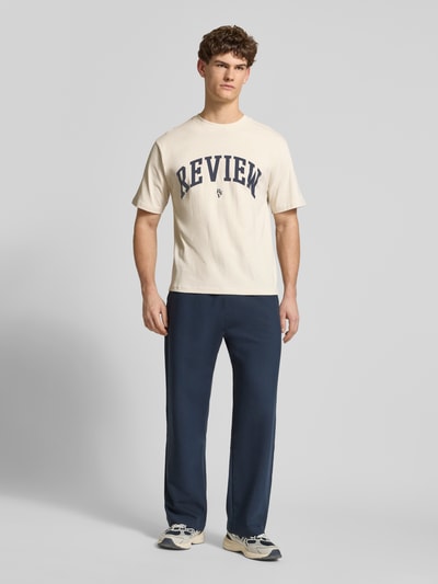 Männermodel trägt T-Shirt mit Label-Print und Rundhalsausschnitt von REVIEW in Beige und Dunkelblau, reine Baumwolle, Ganzkörperansicht von vorne