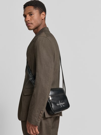 Männermodel trägt dunkle CK Calvin Klein Herren Umhängetasche mit Logo-Prägung, Silberdetails, glatte Oberfläche, Ganzkörperansicht Rückansicht.