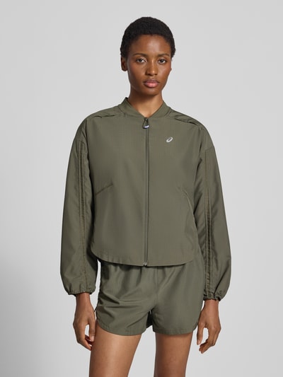 Kobiece model prezentuje kurtka treningowa z elastyczną mieszanką materiałów model 'NAGINO' od ASICS TIGER, odblaskowe detale, luźny krój, stójka, zamek błyskawiczny, przegródka zapinana na zamek błyskawiczny z tyłu, elastyczne mankiety, długość tyłu w rozmiarze S: 51 cm, szerokość ramion w rozmiarze S: 53 cm, szerokość klatki piersiowej w rozmiarze S: 50 cm, długość rękawów w rozmiarze S: 53 cm, oliwkowy kolor, gładka struktura, matowe wykończenie, całkowite ujęcie z przodu.