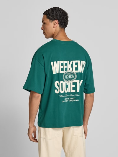Männermodel trägt Oversized T-Shirt mit rückseitigem Print von REVIEW in Grün, Bio-Baumwoll-Mix, Oversized-Fit, Logo-Print, Ganzkörperansicht Rückansicht.