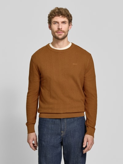 Männermodel trägt Regular Fit Pullover aus Baumwoll-Woll-Mix von GANT in Braun mit Fischgrätmuster, blaue Jeans, Ganzkörperansicht von vorne. Kontext: Herren Pullover von GANT