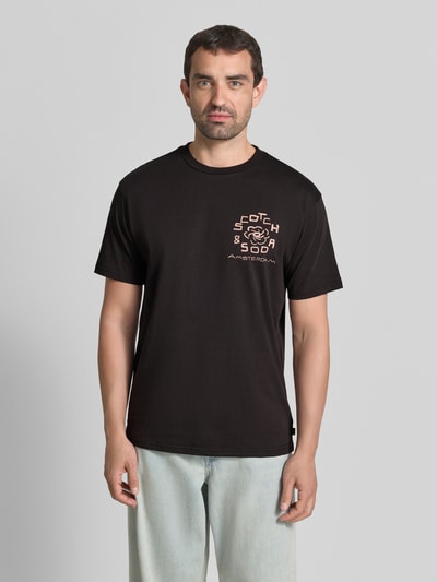 Męski model nosi T-Shirt mit Label-Print und Rundhalsausschnitt von Scotch & Soda z czarnej bawełny, krój regular fit, długość tyłu 72 cm, Ganzkörperansicht von vorne.