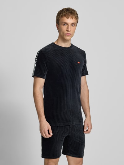 Männermodel trägt T-Shirt mit Logo Modell 'MADORI' von Ellesse aus schwarzem Baumwoll-Mix mit Stretch-Anteil, Regular-Fit, Rundhalsausschnitt, Logo, Rückenlänge bei Größe M 66 cm, feine Rippenstruktur, Ganzkörperansicht von vorne.