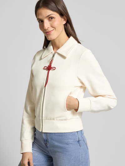 Modelka kobieca nosi Slim Fit Sweatjacke aus reiner Baumwolle Modell 'EROICO' od MAX&Co., kolor jednolity, z zamkiem, krój slim fit, ozdobny ścieg i metaliczne akcenty, długi rękaw, stójka koszulowa, kieszenie boczne, detal z kokardą przy zamku, dzianina, bawełna, widok całej sylwetki z przodu.
