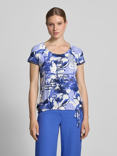 Frauenmodel trägt Damen T-Shirt aus Viskose-Mix mit Kordelzug am Saum von Betty Barclay, blau gemustert mit Statement-Print, Strasssteinbesatz, Regular-Fit, Stretch-Anteil, Rundhalsausschnitt, Kordelzug und Schnürung am Saum, Rückenlänge bei Größe 36: 60 cm, Ganzkörperansicht von vorne.