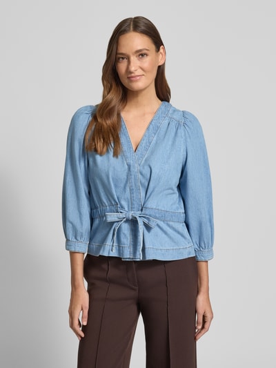 Frauenmodel trägt Relaxed Fit Bluse in Denim-Optik Modell 'Lavina' von Bash aus reiner Baumwolle, 3/4-Arm, taillierte Passform, V-Ausschnitt, Wickel-Optik, blaue und braune Farbtöne, strukturierte Oberfläche, zusammengefasste Ärmelabschlüsse, Ganzkörperansicht von vorne.