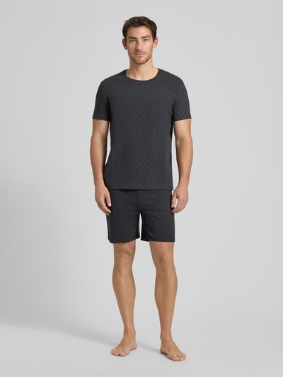 Męski model nosi Shorts aus Baumwolle mit Logo-Muster od JOOP! Collection, czarny kolor, wzór z logo, krój regular fit, szerokość w rozmiarze M: 42 cm, długość wewnętrzna nogawki: 18 cm, bawełna, elastyczny pas. Ganzkörperansicht z przodu.