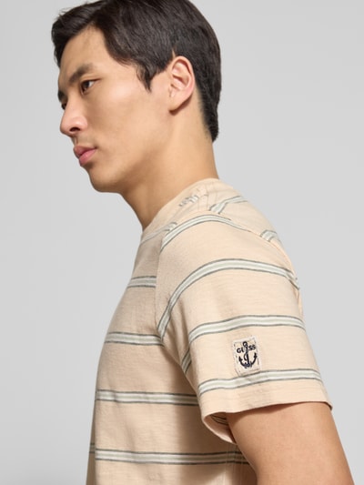 Männermodel trägt T-Shirt mit Rundhalsausschnitt und Label-Patch von Guess in Beige mit Streifen, reine Baumwolle, Nahaufnahme Oberkörper.