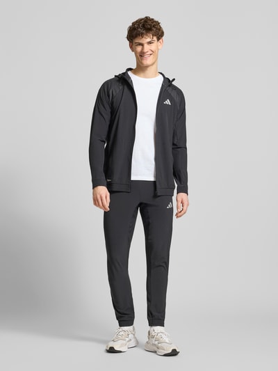 Männermodel trägt schwarze Sweatjacke mit Stretch-Anteil Modell 'TECH' von adidas Training, gerade Passform, Ganzkörperansicht von vorne.