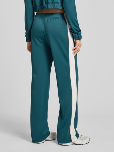 Frauenmodel trägt Sweatpants mit elastischem Bund und Label-Stitching von New Balance in Petrol mit weißem Streifen, Relaxed-Fit, Eingrifftaschen, Innenbeinlänge 80 cm bei Größe S, Ganzkörperansicht Rückansicht.