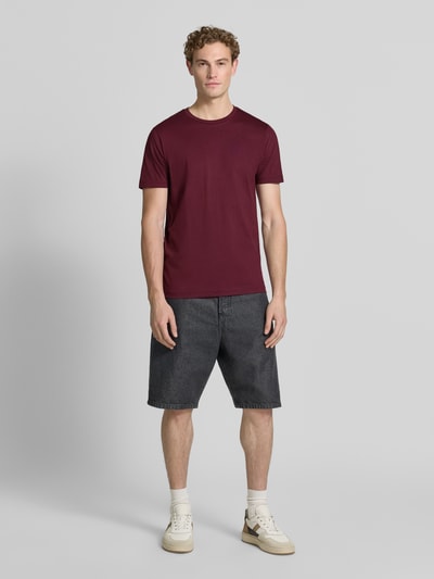 Męski model nosi T-Shirt aus Baumwolle mit Rundhalsausschnitt von Christian Berg Men, krój slim fit, okrągły dekolt, długość tyłu 73 cm w rozmiarze M, kolor bordowy, tekstura gładka, ciemne szorty, jasne buty, całkowity widok z przodu.
