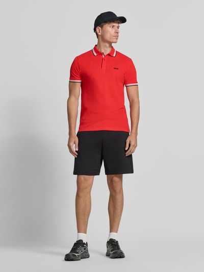 Mężczyzna nosi koszulkę polo Regular Fit Poloshirt aus reiner Baumwolle model 'PADDY' od BOSS Green, czerwona, czarne szorty, całkowity widok z przodu.