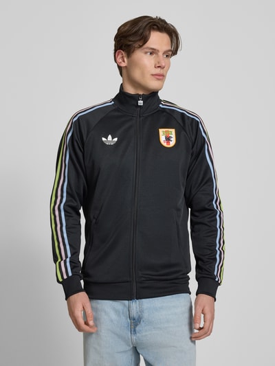 Männermodel trägt Regular Fit Trainingsjacke mit JFA-Applikation von adidas Originals in Optik: Unifarben mit Kontraststreifen, Logo-Stitching, Kragenform: Stehkragen, Ärmelform: Langarm, Ärmelabschluss: Gerippt, Taschen: Seitliche Eingrifftaschen, Verschluss: Reißverschluss. Ganzkörperansicht von vorne.