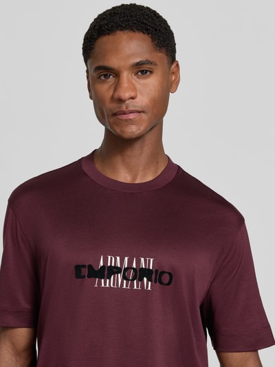 Männermodel trägt T-Shirt mit Label-Print von Emporio Armani in Bordeaux, glatte Textur, gerader Schnitt, Rundhalsausschnitt, Nahaufnahme Oberkörper.