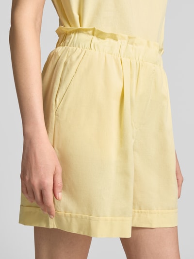 Kobieta nosi Regular Fit bermudy z bawełny i lnu model 'SELMA' od Vero Moda, efekt jednolity kolor, popelina, wpuszczane kieszenie, zapięcie na haftkę i ukryty zamek błyskawiczny, szlufki na pasek, wykończenie materiału: mieszanka bawełny i lnu, kolor jasnożółty, detal: gładka tekstura, zdjęcie z przodu, zbliżenie na dolną część tułowia.