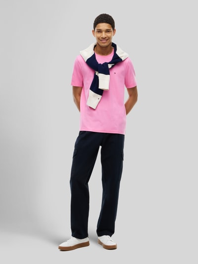 Männermodel trägt Regular Fit T-Shirt aus reiner Baumwolle von Tommy Hilfiger in Rosa mit dunkler Hose, Ganzkörperansicht von vorne.