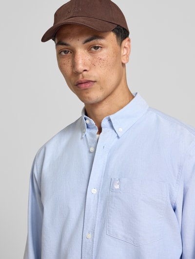 Männermodel trägt Regular Fit Freizeithemd mit Brusttasche und Logo-Stitching von Carhartt Work In Progress in Hellblau aus Reine Baumwolle, Button-Down-Kragen, Knopfleiste, Sportmanschetten, abgerundeter Saum, Nahaufnahme Oberkörper.