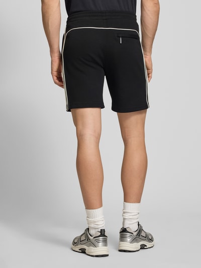 Męski model nosi szorty z wyhaftowanym logo model 'EGEO' od SERGIO TACCHINI, mieszanka bawełny z dodatkiem streczu, luźny krój, elastyczny pas z trokiem, wpuszczane kieszenie, szerokość w rozmiarze M: 40 cm, długość wewnętrzna nogawki: 20 cm, czarne z białymi lamówkami, tekstura gładka, widok całej sylwetki z tyłu.